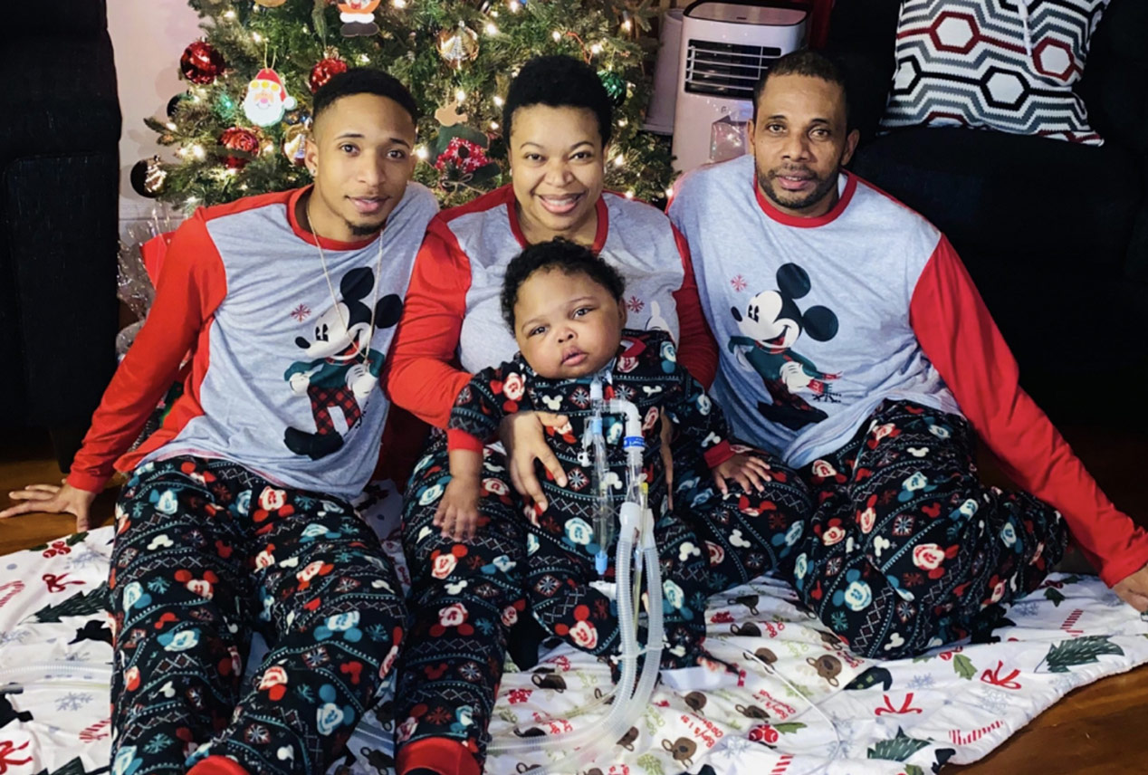 Three parents in matching pajamas in front of a Christmas tree with a young child in the mothers lap