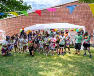 A large group of campers in tie-dyed shirts posing in the grass under a tent on a sunny day